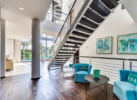 Modern loft interior with floating staircase, turquoise seating, wooden floors, and large windows overlooking palm trees