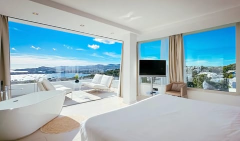 Modern hotel room with white bed, freestanding bathtub, floor-to-ceiling windows overlooking cityscape