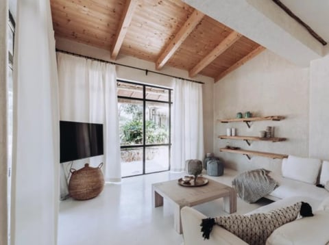 Modern minimalist living room with wooden ceiling beams, white walls, TV, floating shelves, and neutral-toned furniture