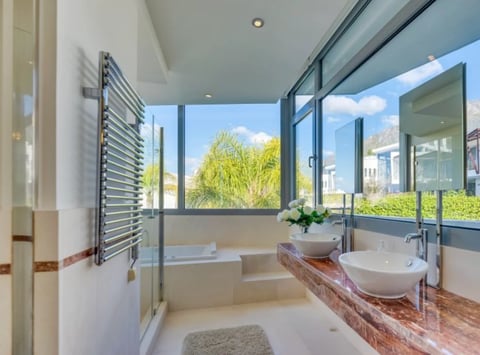 Modern bathroom with large windows, freestanding bathtub, dual vessel sinks on wooden counter, and city skyline view