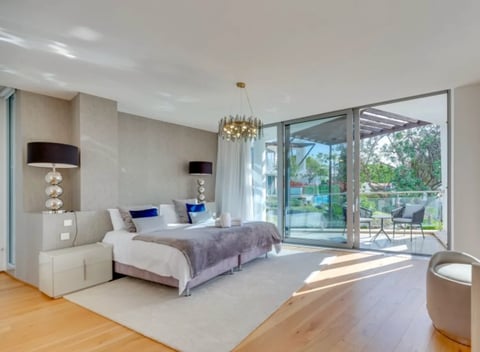 Modern bedroom with large glass doors opening to a garden, featuring a bed with blue pillows, cream furnishings, and natural sunlight