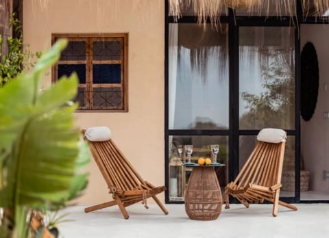 Outdoor patio with two wooden lounge chairs, woven table, and tropical plants against a peach-colored building with thatched roof details