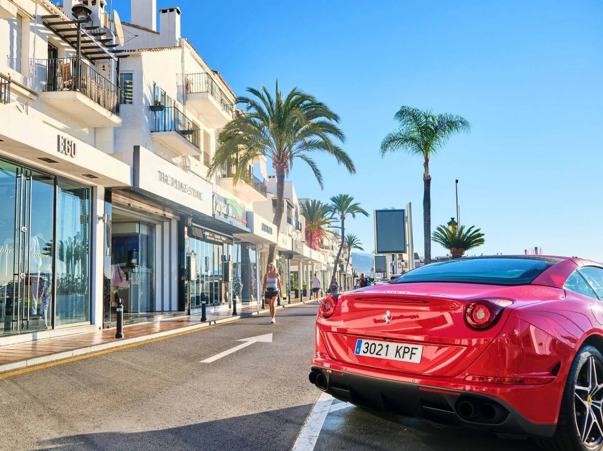 Red sports car parked on a palm-lined promenade with shops and white buildings under clear blue sky