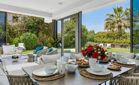 Modern dining room with large windows overlooking a garden, featuring a table set for dining with red flowers and views of palm trees and lawn outside
