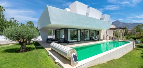 Modern white geometric house with bright green swimming pool, manicured lawn, and mountain backdrop