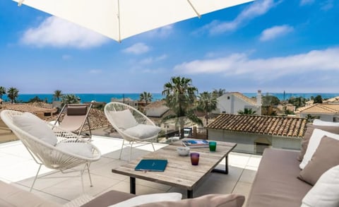 Modern rooftop terrace with white chairs, wooden table, and ocean view under blue sky with palm trees