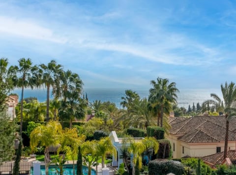 Coastal Mediterranean landscape with palm trees, lush vegetation, tile-roof villa, and ocean view under blue sky