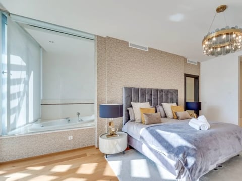 Modern luxury bedroom with en-suite bathroom, featuring a gray upholstered bed, sunlit space with wooden floors, and contemporary pendant lighting