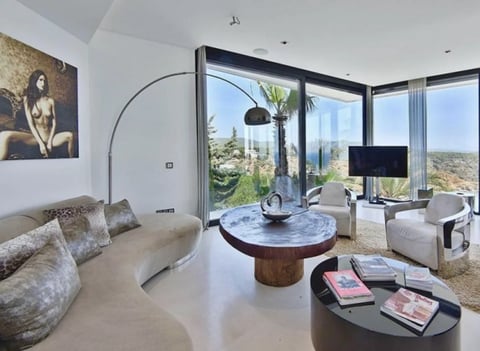 Modern luxury living room with floor-to-ceiling windows, arc lamp, round marble table, beige seating, and panoramic city views