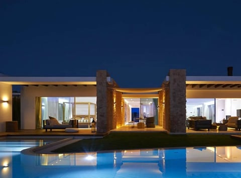 Modern luxury home at dusk with illuminated interior, swimming pool, and two prominent stone columns reflected in water