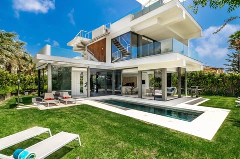 Modern two-story white villa with pool, lounge chairs on manicured lawn, blue sky, surrounded by palm trees