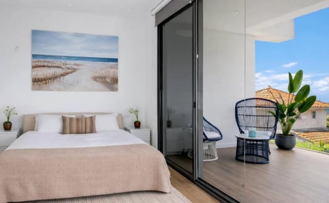 Modern bedroom with bed, beach artwork, and open sliding glass doors leading to a balcony with seating and potted plant