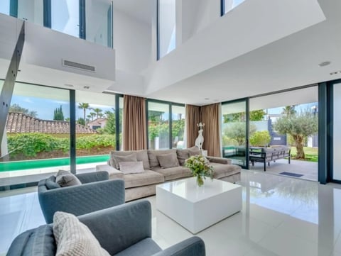 Modern open-plan living room with white cubic coffee table, beige and gray seating, large windows overlooking pool and garden landscape