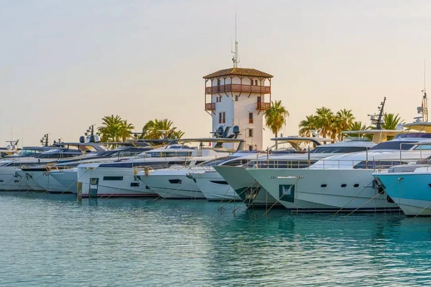 Row of turquoise luxury yachts moored in clear blue water with a historic lighthouse tower and palm trees in background