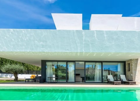 Modern minimalist house with white exterior, large glass doors, olive tree, and bright turquoise pool under blue sky
