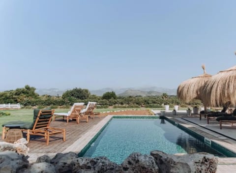 Luxurious resort pool with wooden loungers, thatched roof structure, and mountains visible in the background under clear sky