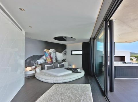 Modern luxury bedroom interior with round bed, dark headboard wall, large windows, and open door to outdoor patio deck