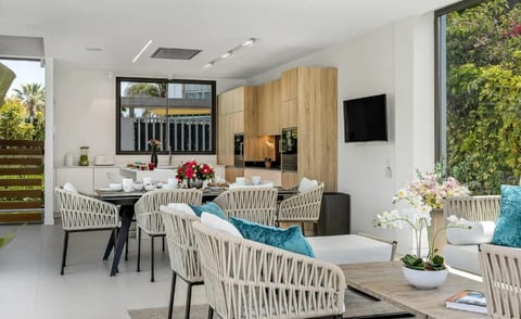 Modern open-plan living space with rope-woven dining chairs, kitchen area, and natural outdoor views through large windows