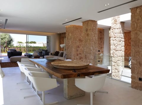 Modern open-plan living space with wood dining table, white chairs, cork accent columns, and floor-to-ceiling windows overlooking outdoor areas