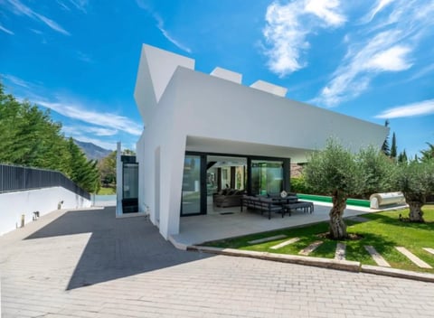 Modern white cubic house with large glass doors, patio seating, olive tree, and manicured lawn under blue sky