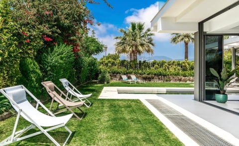 Luxury modern home patio with lounge chairs on manicured lawn, palm trees, and blue sky