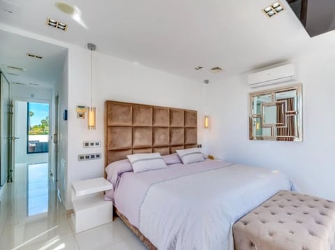 Modern bedroom with queen bed, brown upholstered headboard, white bedding, and beige ottoman, featuring minimalist white walls and natural light