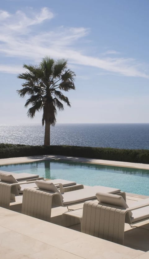 Luxury poolside with lounge chairs, palm tree, and ocean view under clear sky