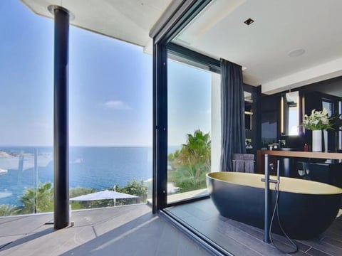 Modern luxury bathroom with black freestanding tub and large glass doors overlooking ocean and coastal city views