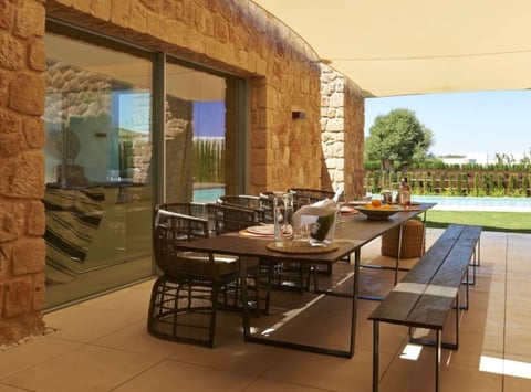 Modern outdoor dining patio with stone walls, wooden table, and pool view in background