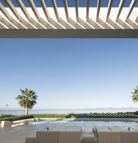 Modern poolside terrace with slatted pergola, lounge chairs, palm trees, and ocean view