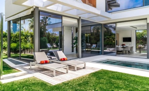 Modern house with glass doors, wooden deck, lounge chairs, swimming pool, and manicured lawn