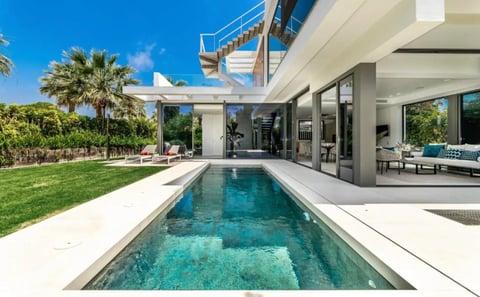 Modern luxury home with narrow rectangular pool, manicured lawn, palm trees, and contemporary architecture with floor-to-ceiling windows
