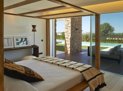 Modern bedroom with wooden beam ceiling and bed overlooking pool and outdoor landscape through floor-to-ceiling glass doors