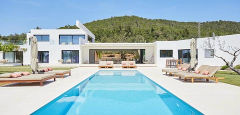 Modern white villa with rectangular swimming pool, sun loungers, and tree-covered hillside
