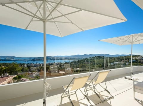Rooftop terrace with white umbrellas and lounge chairs overlooking a coastal bay with mountains