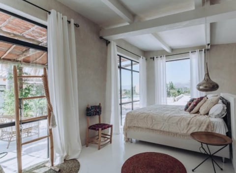 Bright modern bedroom with large windows, white curtains, beige bed with pillows, wooden chair, and reddish-brown round rug