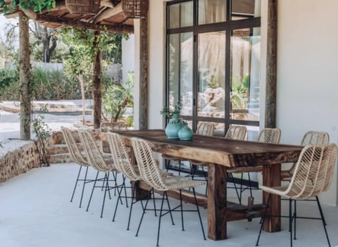 Outdoor dining area with rustic wooden table, woven rattan chairs, and pergola near modern home with large windows
